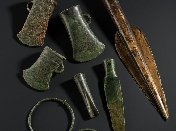 Three axe heads, three spears, and two large rings all made of discoloured bronze sitting on a black background