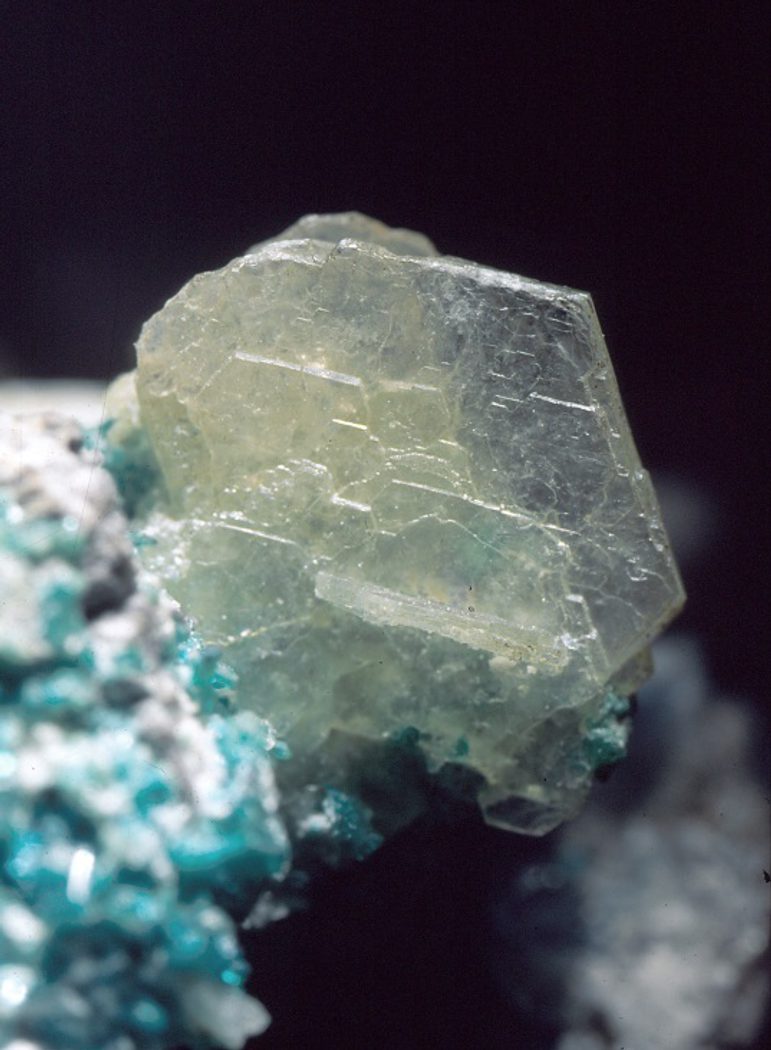 A closeup image of a pale yellow crystal emerging from small blue crystals against a black background