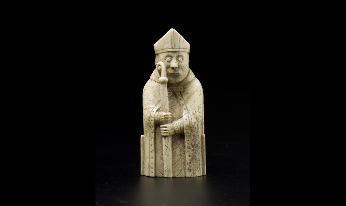 An ivory chess piece of a bishop seated wearing a pointed hat and holding a staff