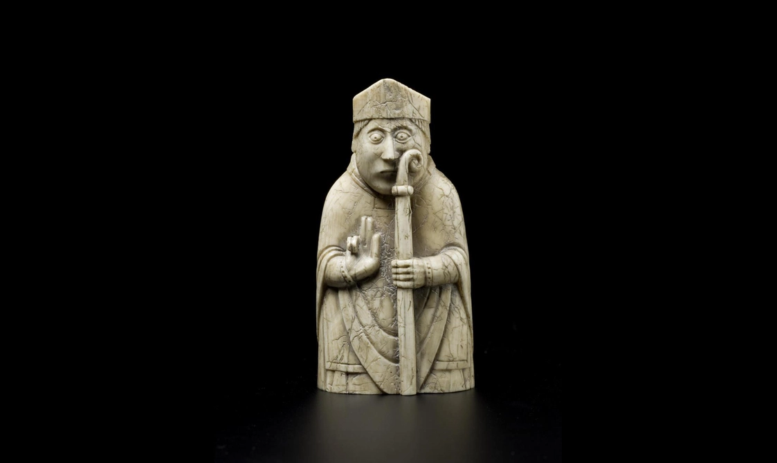 An ivory chess piece of a bishop seated wearing a pointed hat and holding a staff. He is holding up two fingers.