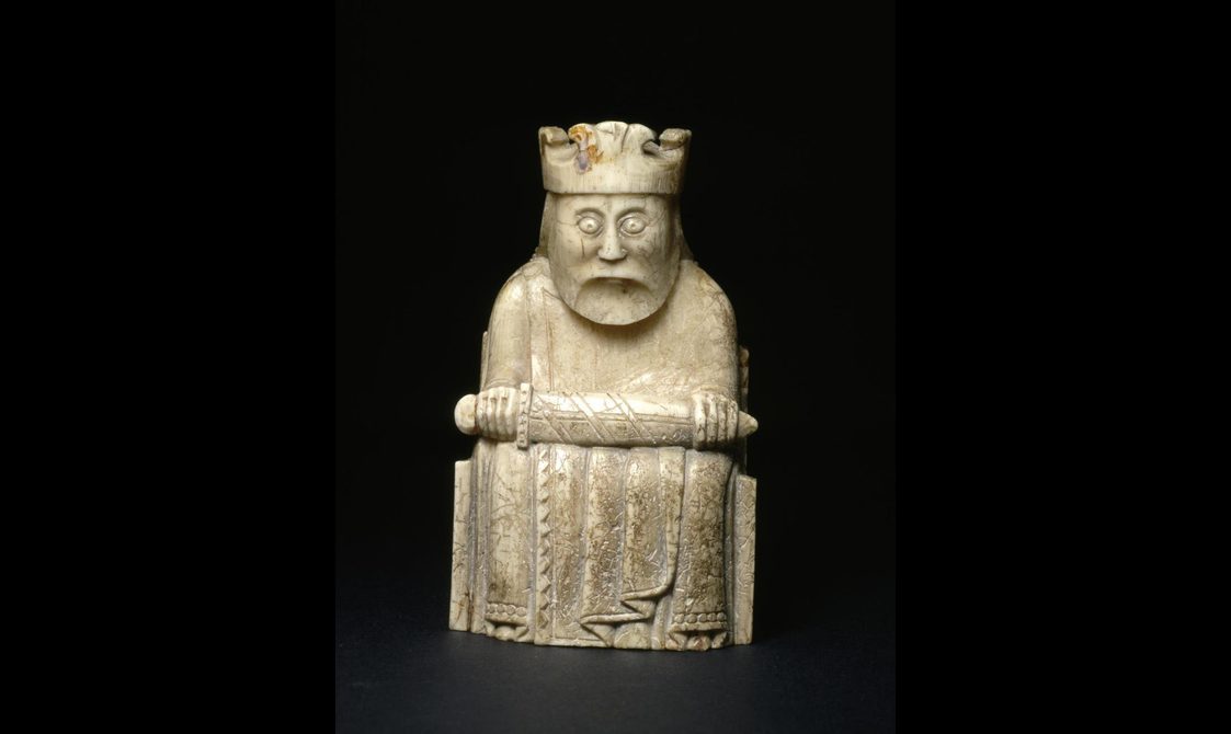 An ivory chess piece of a king seated with a crown on his head with a sword across his lap