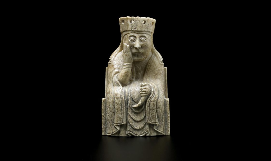 An ivory chess piece of a queen seated with a crown on her head with one hand cradling her face.