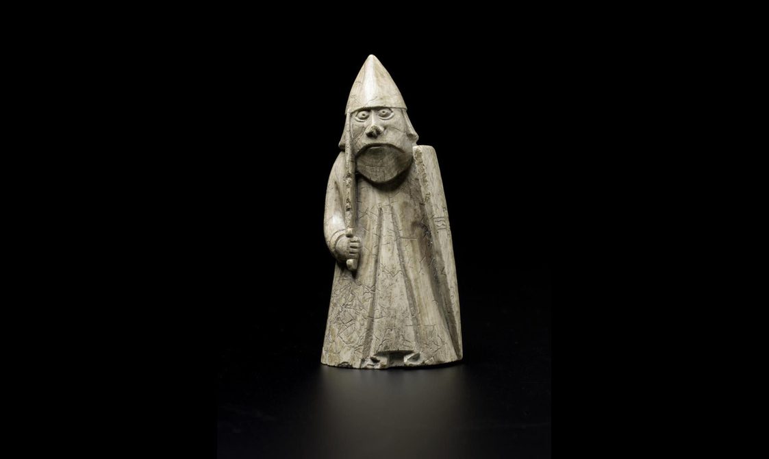 An ivory chess pieces of a figure wearing long robes and a helmet while holding a sword and shield