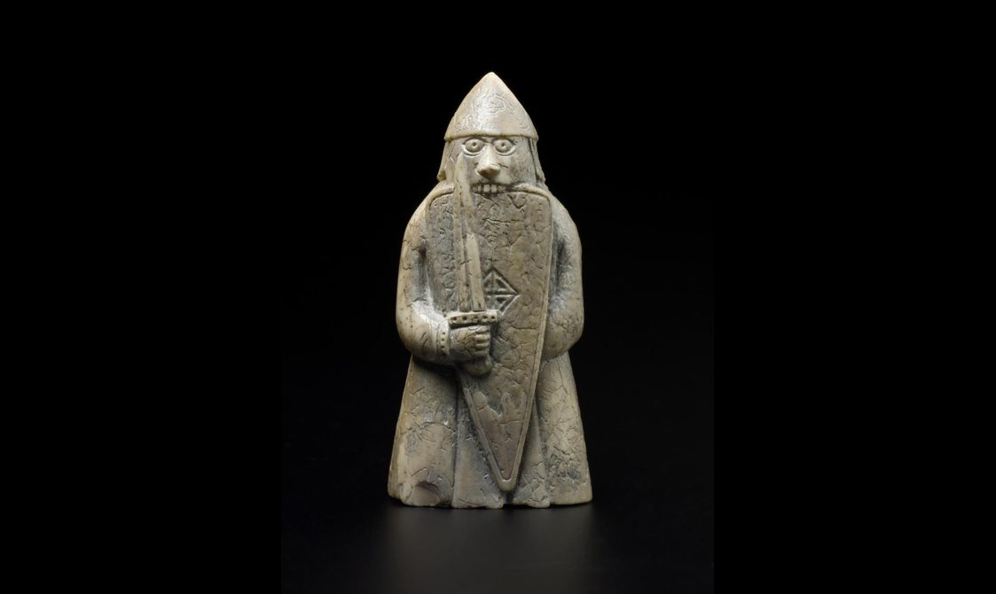 An ivory chess pieces of a figure wearing long robes and a helmet while holding a sword and shield
