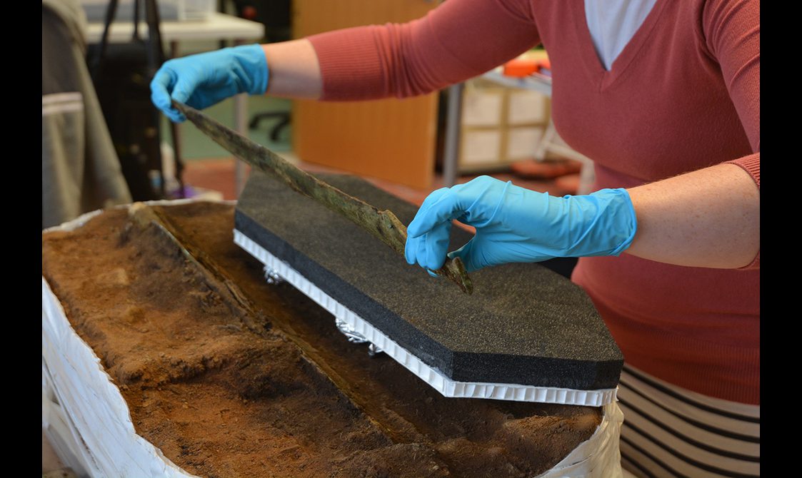 In a conservation lab, Bronze Age sword being held up after being lifted from a block of dirt