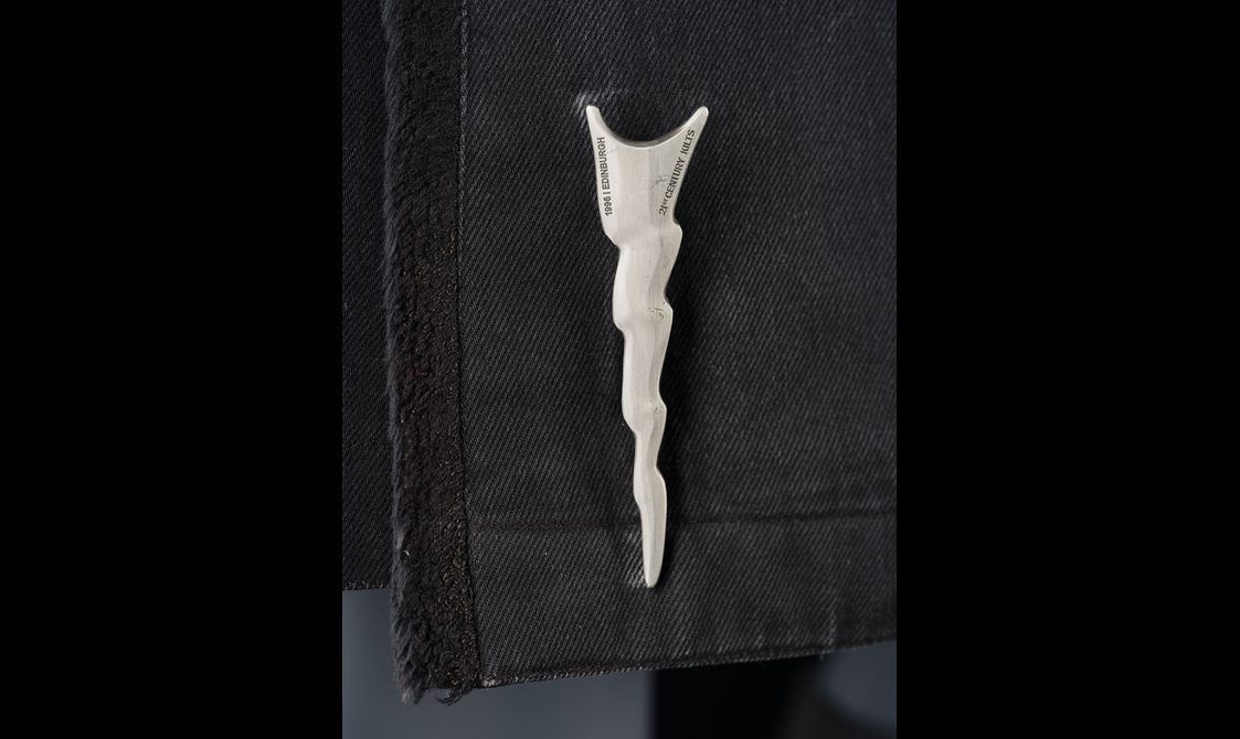 A silver kilt pin in the shape of a lightning bolt attached to a black denim kilt.