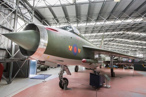 A green military aircraft with a sharp pointed nose in a large aviation hangar.