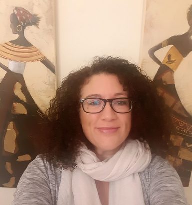 Woman with dark brown hair, grey jumper and white scarf looks forward into the camera with art of two dancing figures behind her.