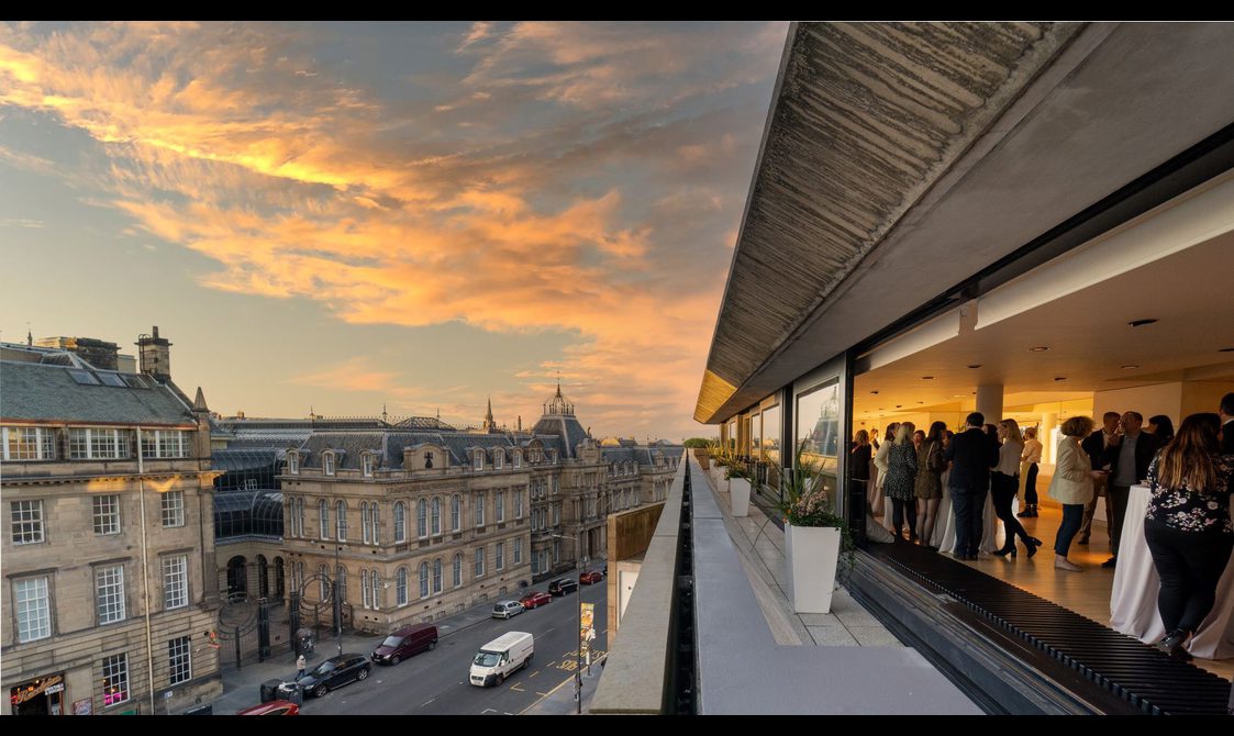 A terrace overlooking the city at sunset. There are open windows revealing people standing around tables with drinks inside.