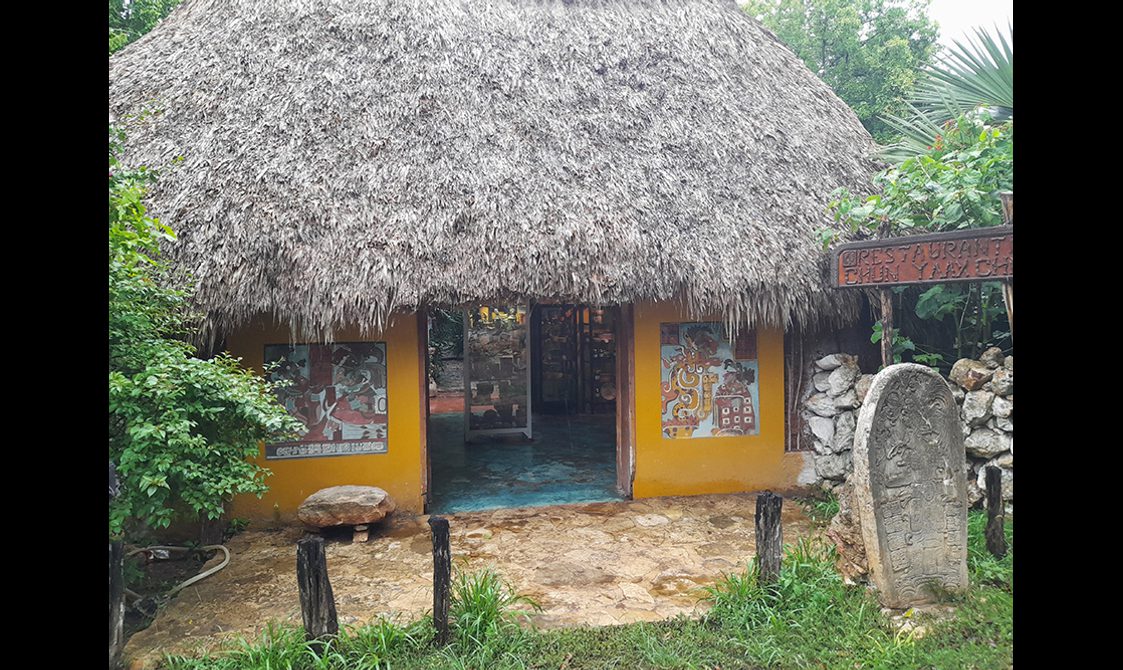 View of the front entrance to Morales's ceramic studio, featuring a yellow building with a thatched roof. Decorative stone carvings and lush greenery surround the entrance.
