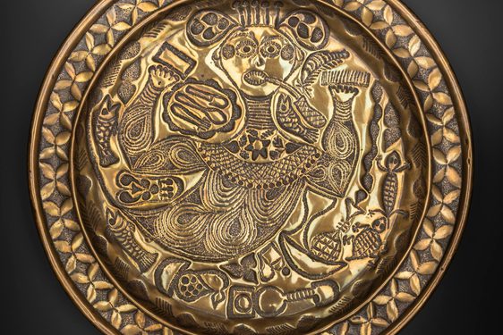 A round gold object with intricate designs engraved including a stylised person at the centre
