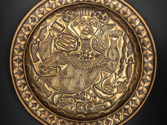 A round gold object with intricate designs engraved including a stylised person at the centre