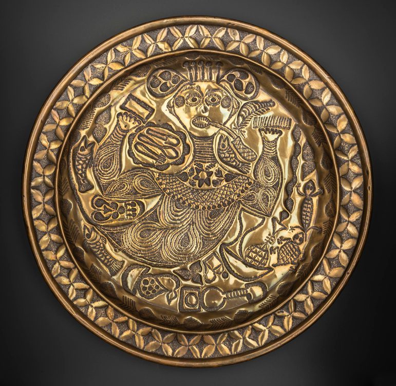 A round gold object with intricate designs engraved including a stylised person at the centre