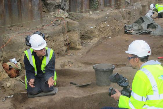 Two archeaologists in high visibility gear in a dig site. One is kneeling down and talking to a video camera, which the other is holding.
