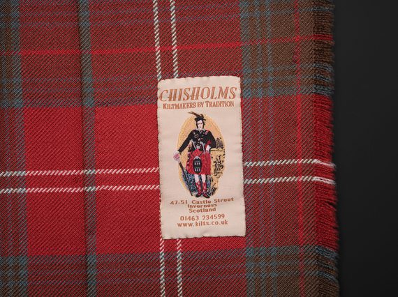 A clothing label with an illustration of a man in a dress kilt and shop contact details fixed to a red tartan kilt.