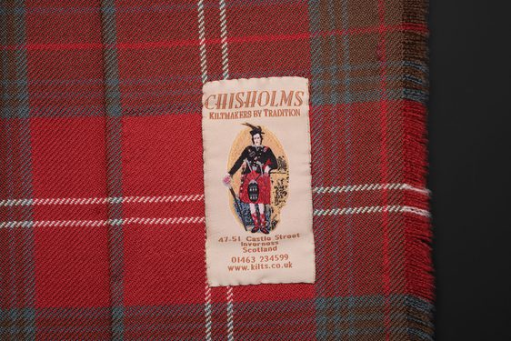 A clothing label with an illustration of a man in a dress kilt and shop contact details fixed to a red tartan kilt.
