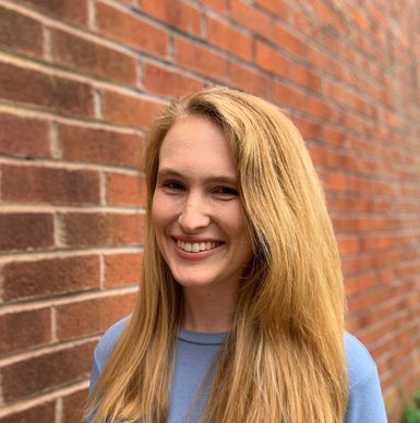 A smiling woman dressed in a blue jumper with blonde hair posing against red brick wall.