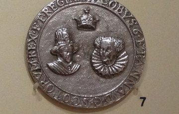 A silver round medal with a king, queen, and a crown with text around the edge.