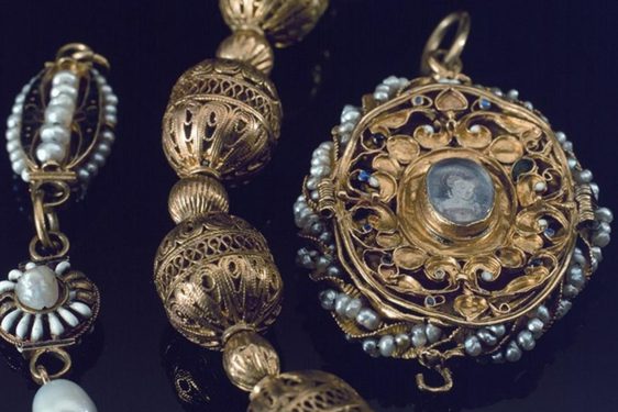 Close up image of three highly decorative pieces of jewelry. One decorated with pearls, the middle piece made out of gold highly decorated baubles, and a third gold locket decorated in pearls and a miniature portrait of a woman.