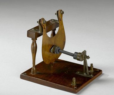 An early model of a microphone which consists of a rectangle wooden stand with a you shaped piece of wood supported by two legs with a metal cylinder going through its centre.