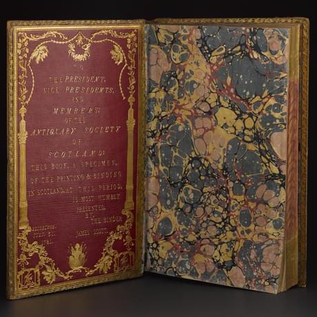 An open book with a red inside cover and gold decoration. The first page is marbled end paper.