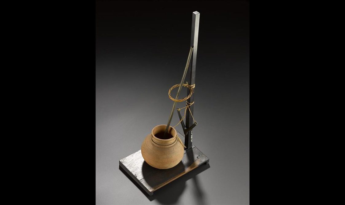 A model of a ceramic pot attached to a long tall rod with string. There is another rod inside the pot that extends to lean against the tall rod.