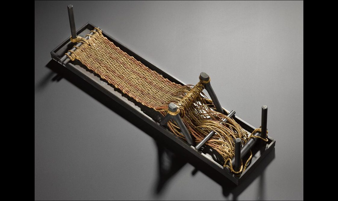 A small rectangular loom with yellow and orange fibres being woven into a textile.