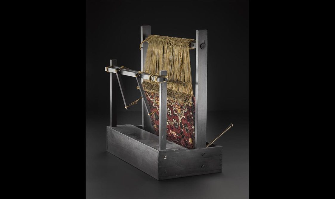 A wooden loom with a red patterned carpet in the process of being made.