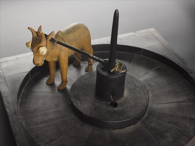A model of a ceramic donkey standing on a wooden wheel with a prong sticking up from the centre and a pole across the donkey's back. The wheel sits on a wooden box.