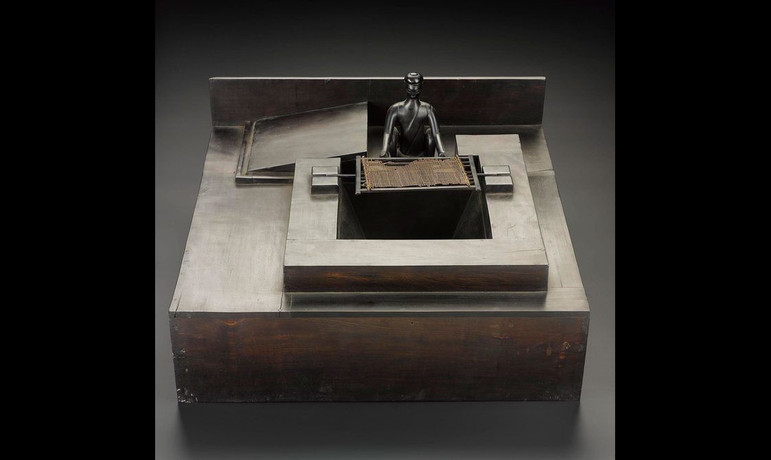 A wooden box with a small metal figure seated with a sieve like rectangle with fabric on top of it.