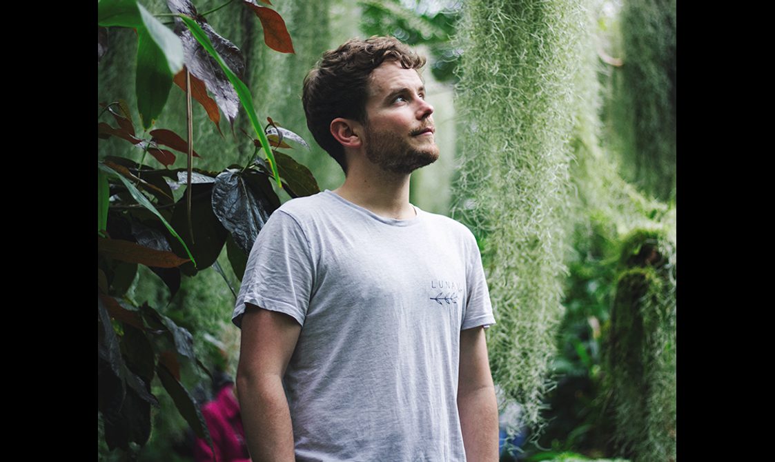 A man looking up, dressed in a white t-shirt with a lush green background.