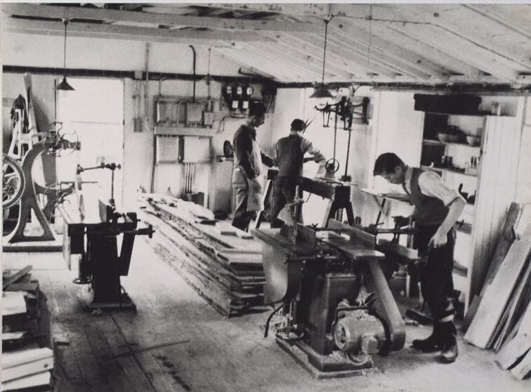 Black and white photograph of three people working at different machines in a shop. There are other unoccupied machines and large sheets of wood stacked up.