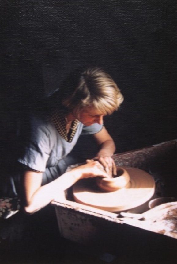 A woman with short blonde hair and a short sleeved top works on a pot on a potter's wheel.