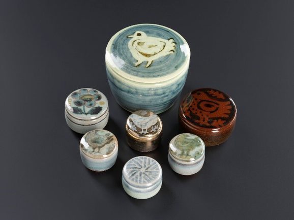 Seven lidded pots of different sizes, glazed in different colours with various patterns on top. Most show different birds, one has a flower, and one a linear pattern.