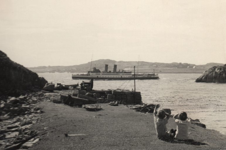 Black and white photo of a ship at sea viewed from a rocky pier. There are two children sitting on the pier looking out at the boat.