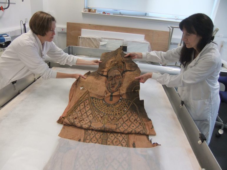 Two women in white lab coats carefully lay a large textile with a painted Egyptian god on its surface on a long white and steel table.