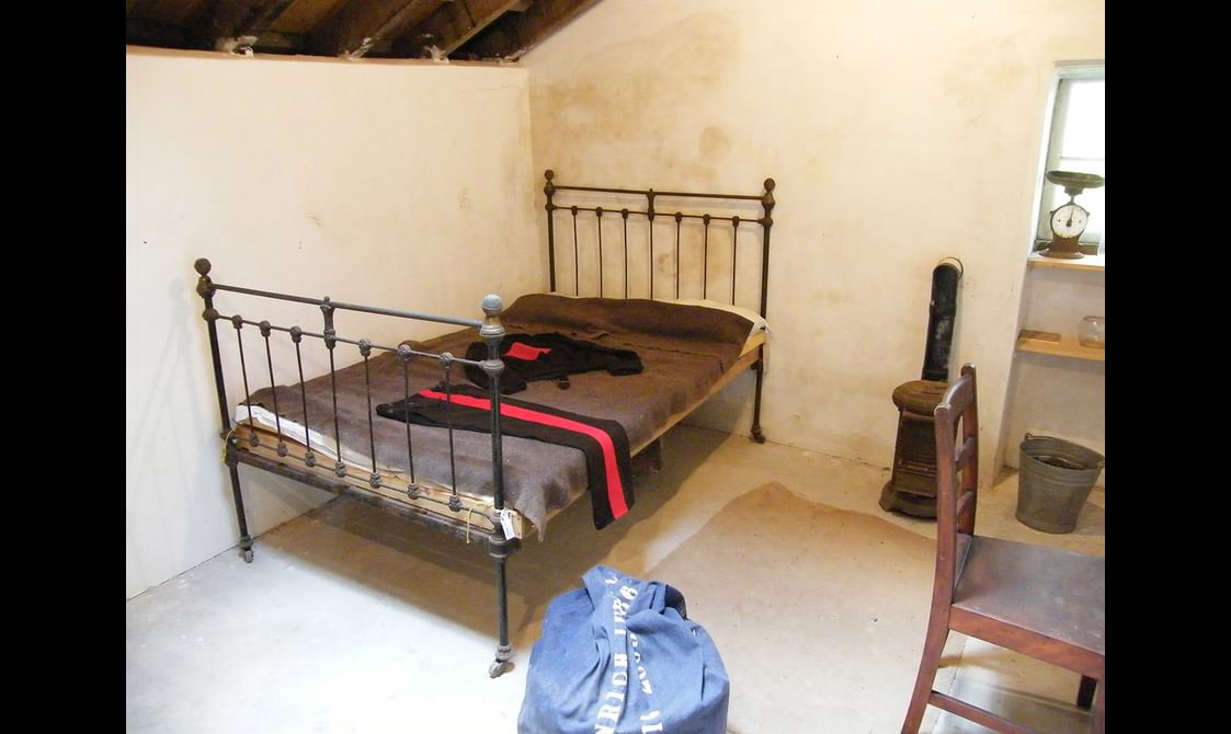 A white walled room with an iron frame bed, with a grey cover and clothing draped over it. There is also a bag, chair, shelf, and bucket.