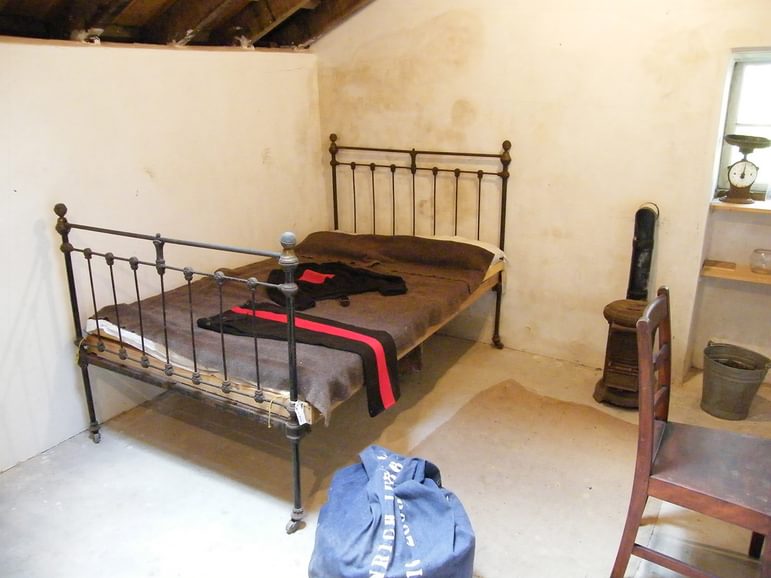 A white walled room with an iron frame bed, with a grey cover and clothing draped over it. There is also a bag, chair, shelf, and bucket.