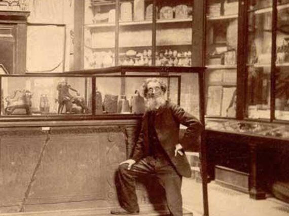 A man photographed standing with one leg raised on a plinth in front of museum case displays. Large display cabinets are visible in the background.