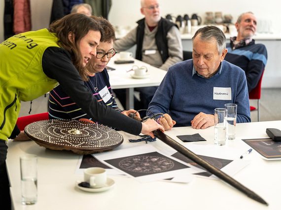 Two people sitting at a table listening to a member of museum staff talk about a sword from the collections.