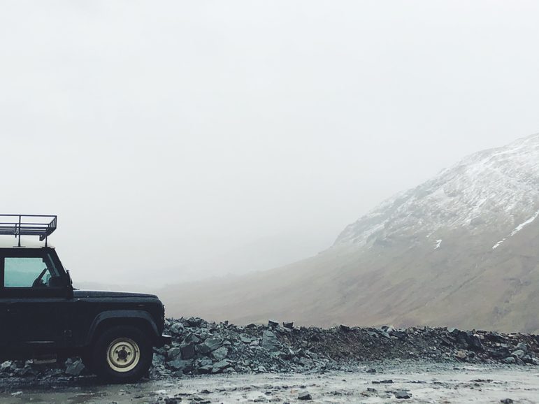 In the foreground, the front end of a Land Rover truck on the left of a rocky-edged road. The background is the side of a snowy mountain and a valley mostly obscured by fog.