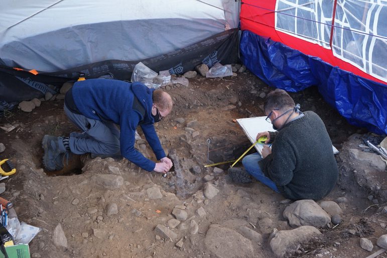Two curators working in an excavation site. One is inspecting something in the ground, whilst the other is taking measurements with a tape measure.