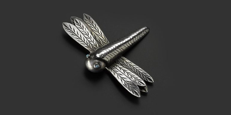 A silver brooch in the shape of a dragonfly with turquoise stones for its eyes. It is laid on a black background.