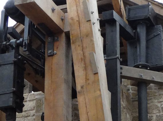 A detail of a large engine made of iron parts and wooden beams attached to a brick building.