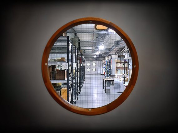 A small circular window, like those you would see in the hull of a ship, gives a glimpse into a museum storage area filled with racks containing boxes and folders.