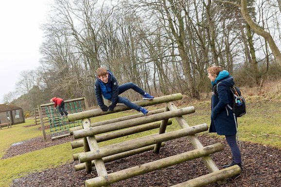 Children play on climbing frames on an assault course while a parent stands by watching.