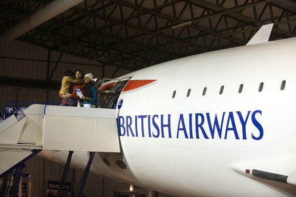Two parents and a child standing at the top of some boarding steps next to a Concorde aeroplane. The plane has "British Airways" written on the side.