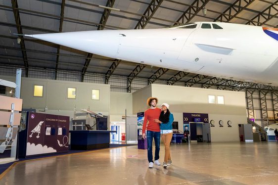 A couple in a museum walkiing under the nose of a Concorde aeroplane.