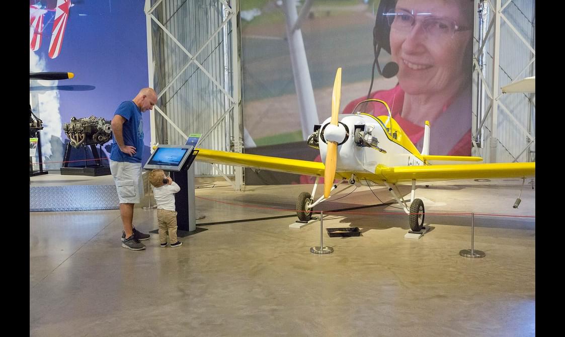 A parent and child interacting with a screen next to a Druine Turbulent aircraft inside the Civil Aviation hangar.
