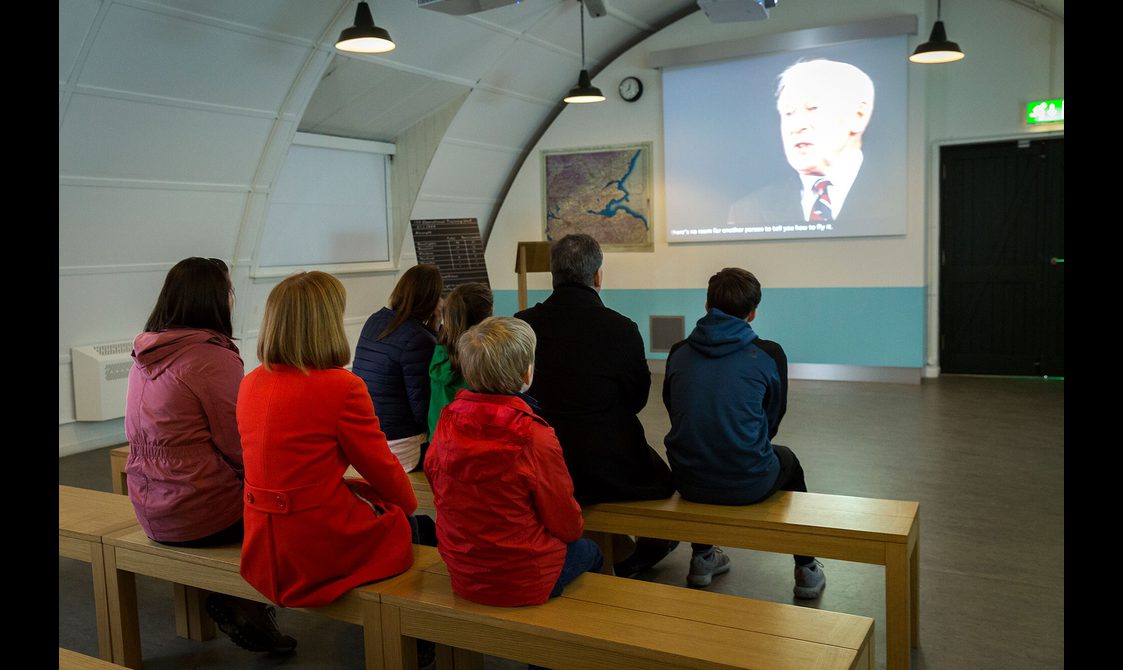 Two rows of visitors sitting watching a film projected in the theatre of the Fortunes of War gallery.